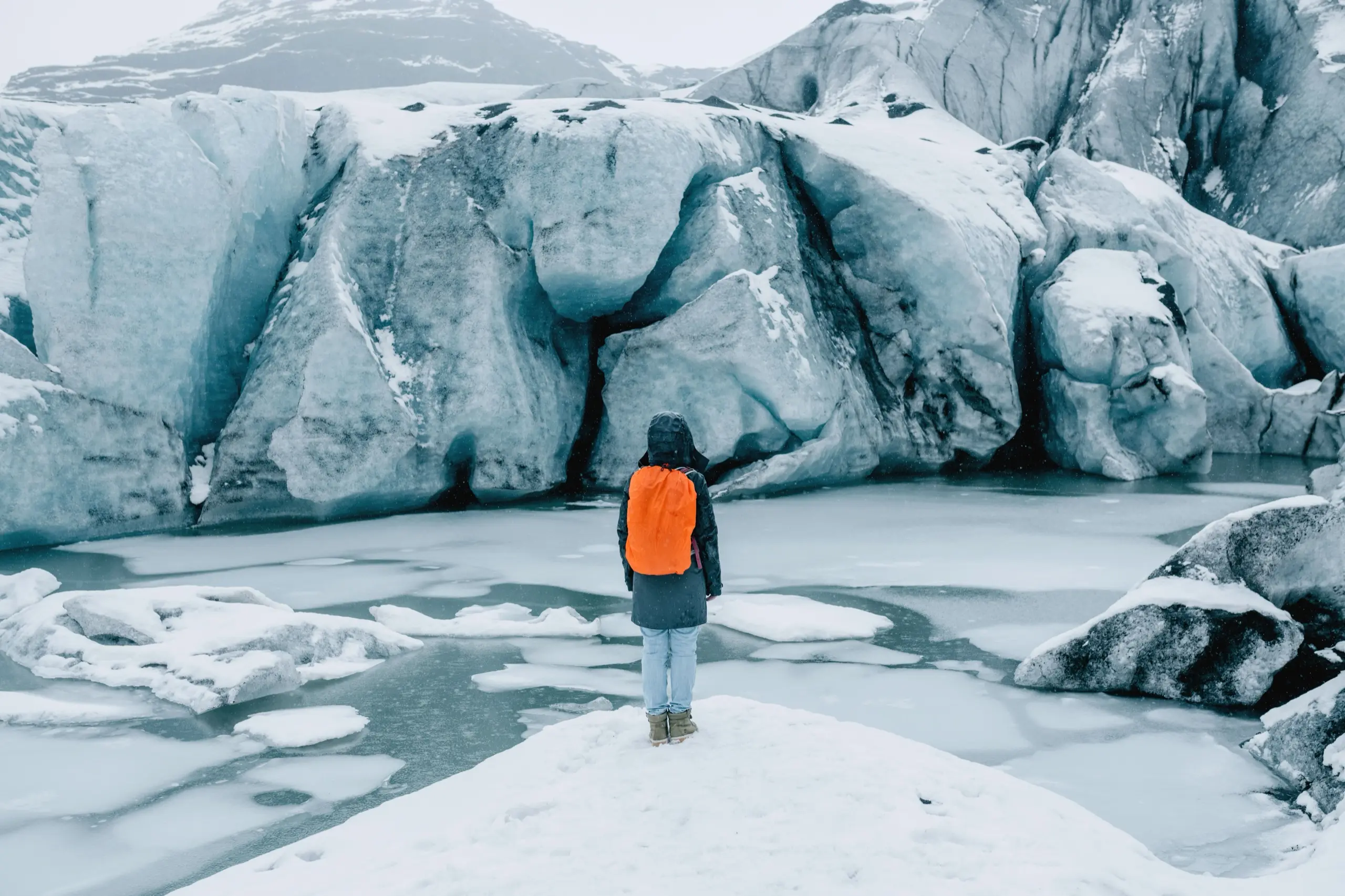 A hiker with an orange backpack standing before a glacier wall and frozen meltwater