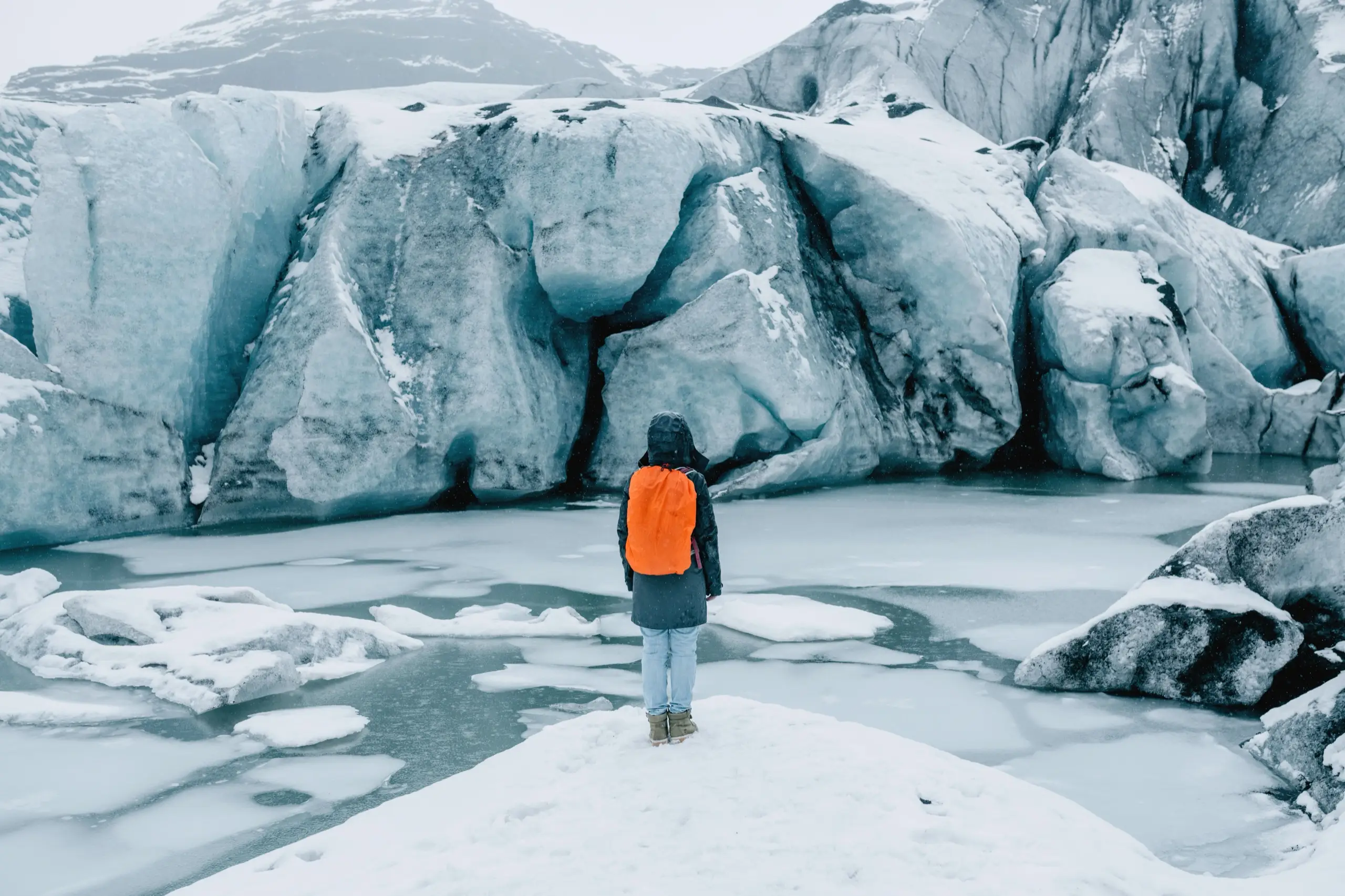 A hiker with an orange backpack standing before a glacier wall and frozen meltwater