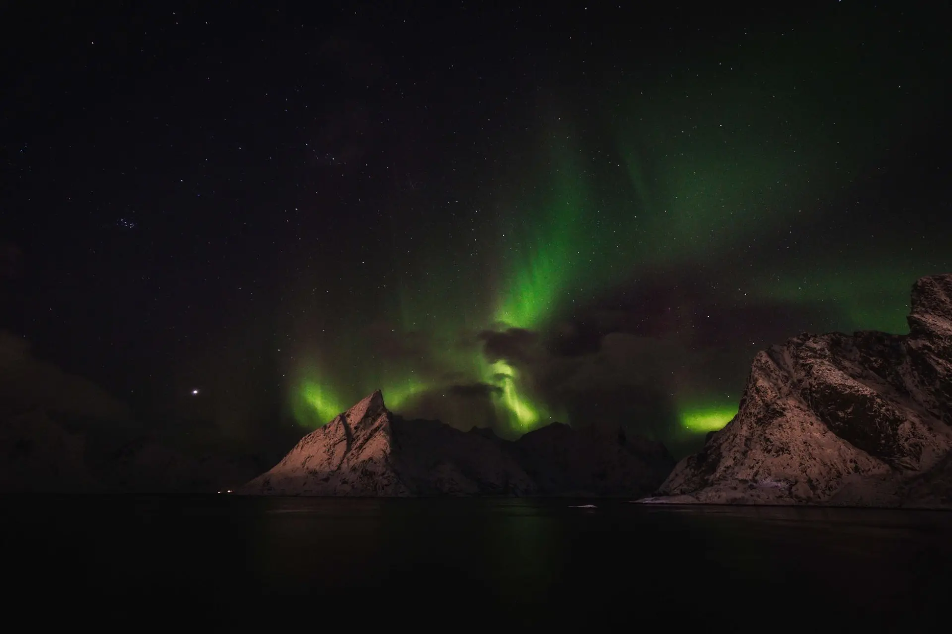 Green northern lights over snow-covered peaks above a dark fjord