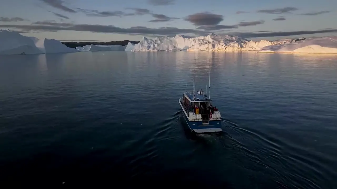 A small tour boat with passengers cruising past glowing icebergs in golden Arctic light